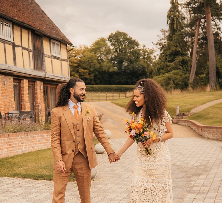 Bride in boho crochet gown walking hand in hand with groom in brown suit with the bride holding a yellow and orange wedding bouquet 