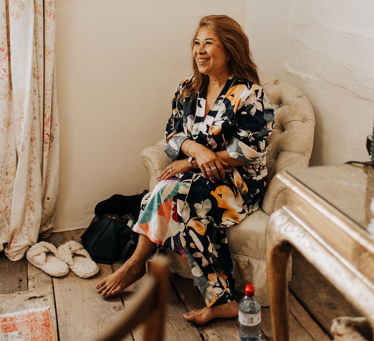 Mother of the bride in a floral robe sits in a chair as she gazes at her daughter on her wedding day 