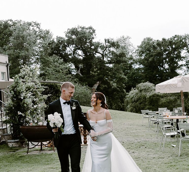 The groom in a black tuxedo walks with the bride in an Aleena Lena wedding dress 