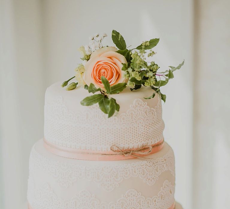 Three tier white iced wedding cake with lace detailing and peach bow ribbon 