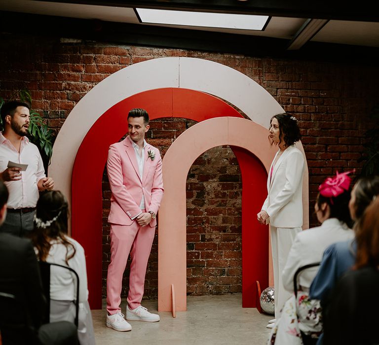 The groom wears a pink wedding suit and the bride wears a white suit as they stand facing each other for their ceremony 