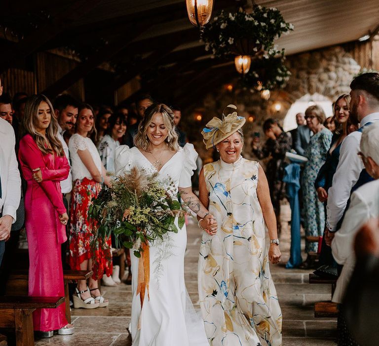 Mother of the bride wearing a beige dress with print and a matching hat walking the bride down the aisle 