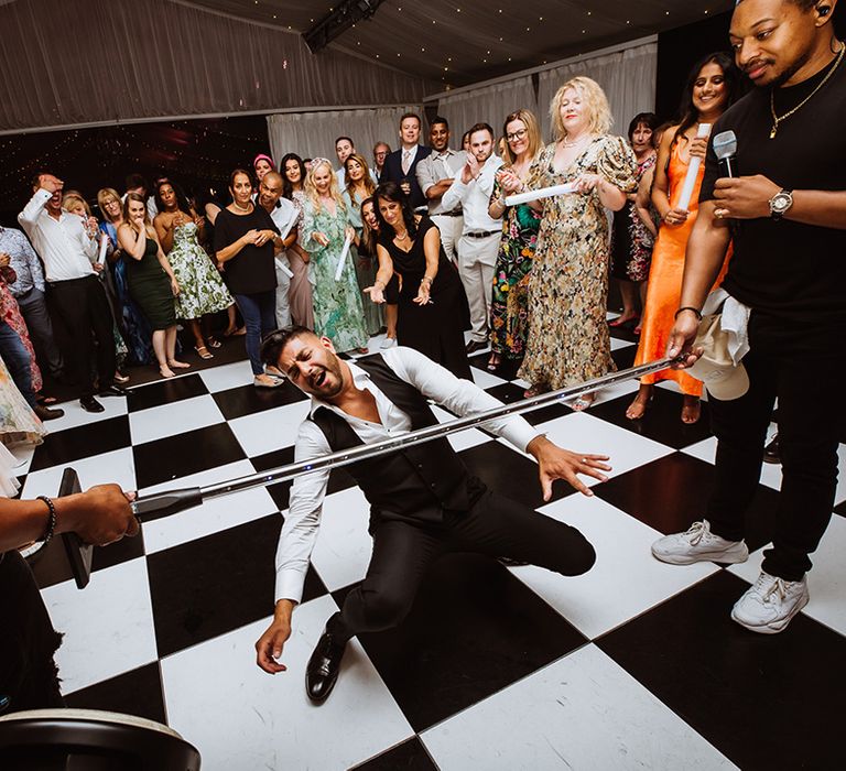 The groom plays a game of limbo at the wedding reception on a black and white checkered floor 