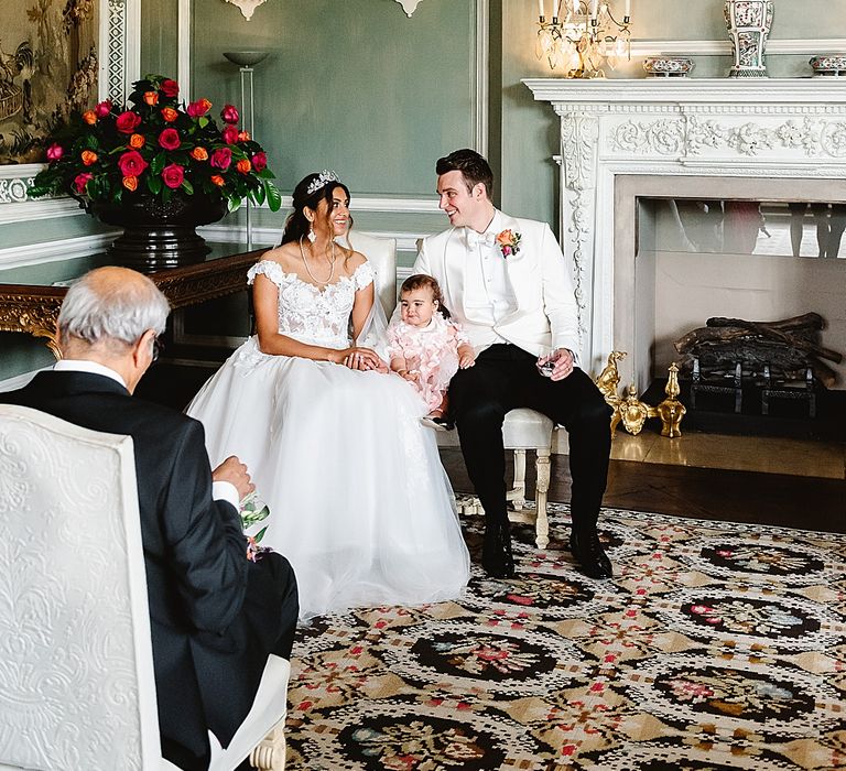 Bride in an off the shoulder wedding dress and groom in a white tuxedo sitting with their daughter during their wedding ceremony 