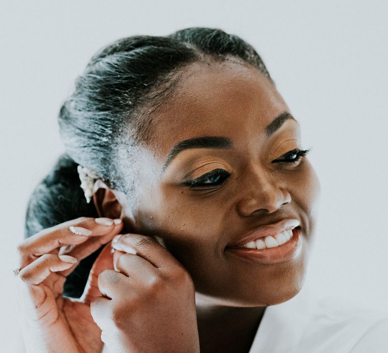 A Black bride outs an earring in as she gets ready for her wedding. She has a low bun in her hair and central parting.