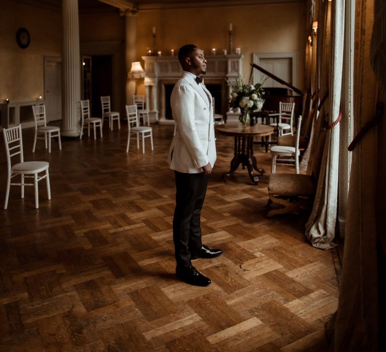 Groom in a white double breasted tuxedo jacket and black bow tie waiting for his bride to arrive at their first look. 