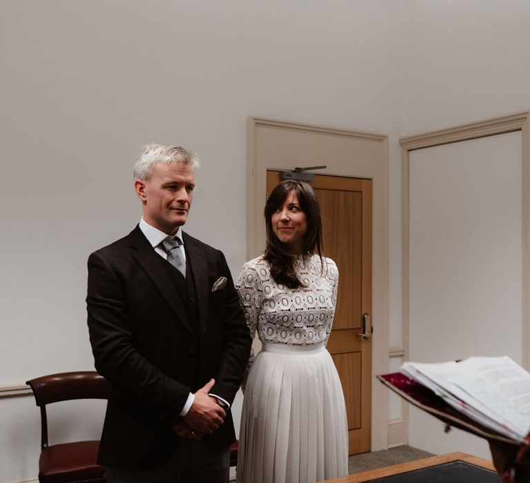 Bride in Self Portrait wedding dress looks sideways at groom in brown woollen blazer and waistcoat whilst getting married at Bristol Registry Office