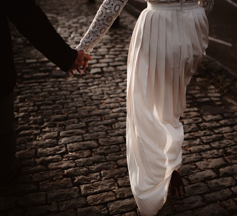 Bride in long sleeved lace top Self Portrait wedding dress with pleated skirt holding hands with groom in brown woollen blazer walking through cobbled street in Bristol 