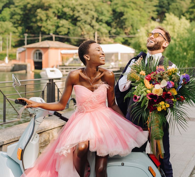 Black bride sits on Vesper in pink wedding gown whilst laughing with groom who holds colourful bouquet 