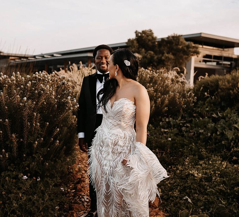 Bride in a strapless Biji La Maison de Couture wedding dress with feather design leading her groom in a black tuxedo through the vineyards