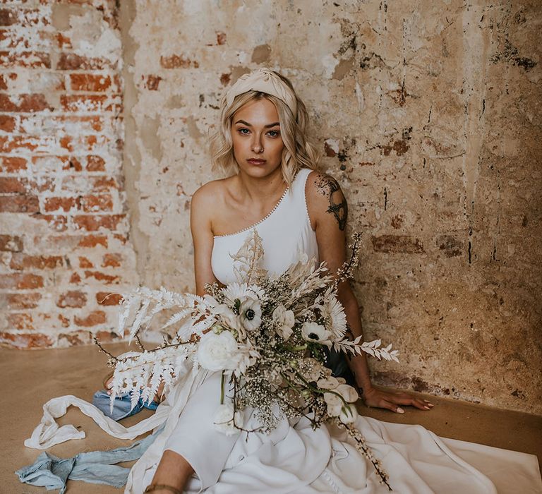 Bride in a one shoulder wedding dress with buttons on the back wearing a headband and holding a white flower bouquet 