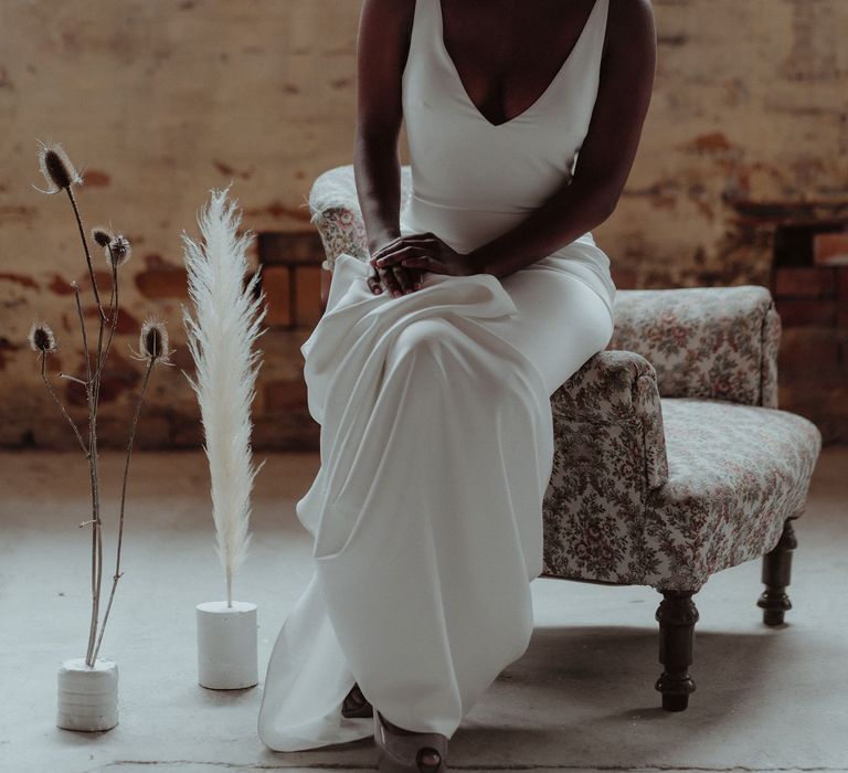 A Black bride sits on the arm of a chair in a low cut, sleek white wedding dress. Her hands sit in her lap.