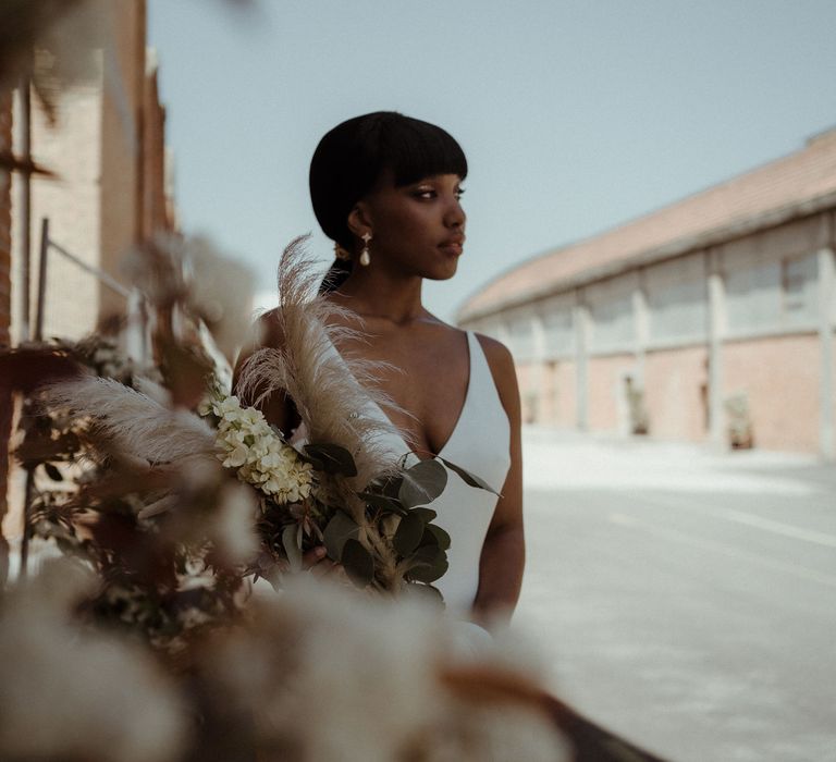 A bride holds a dried flower and grass bouquet. She wears a low v neck wedding dress. 