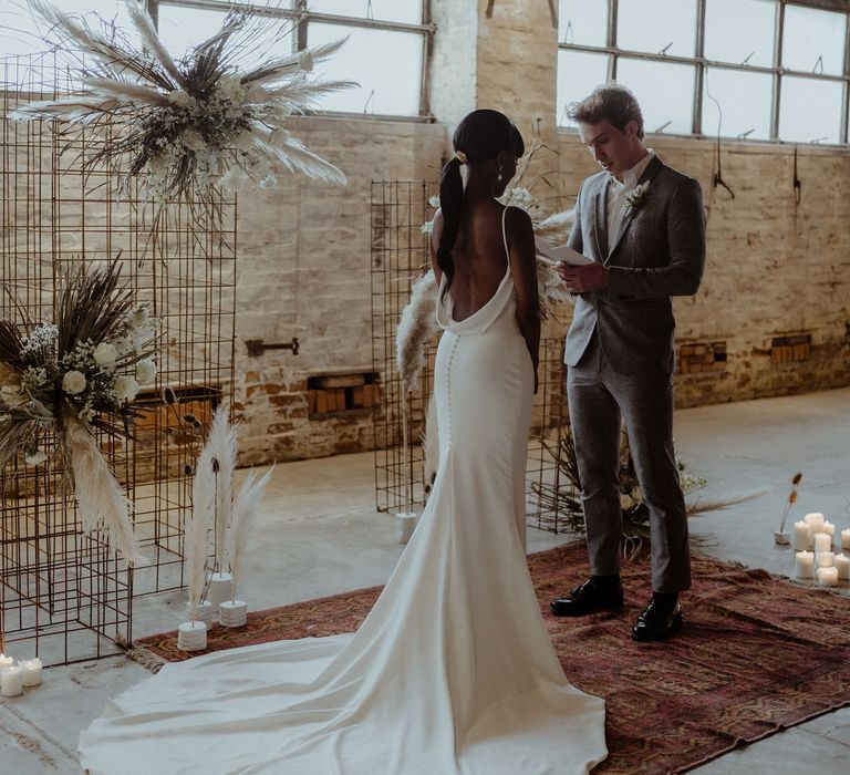 A bride and groom stand in front of a metal frame structure and he reads his vows from a book. She wears a long sleek white dress with a train and he wears a grey suit. They stand on a rug,