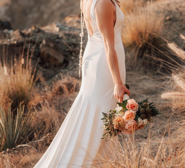 Bride looks down as she holds peach bouquet with green foliage during golden hour