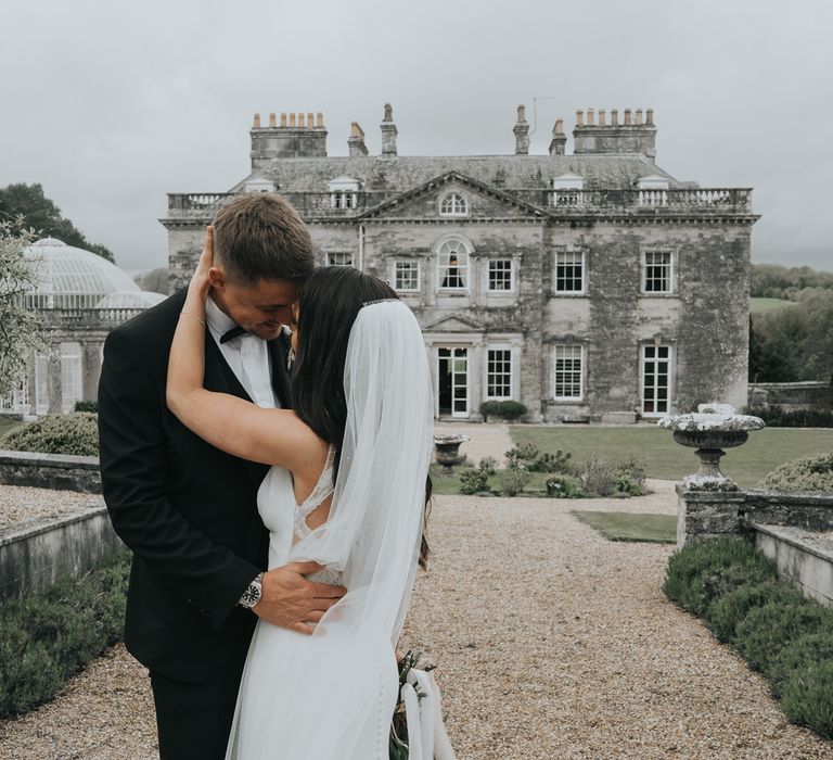 Bride in white Pronovias wedding dress and veil holds grooms head as he wraps arms around her as they stand in front of Came House Dorset after their wedding