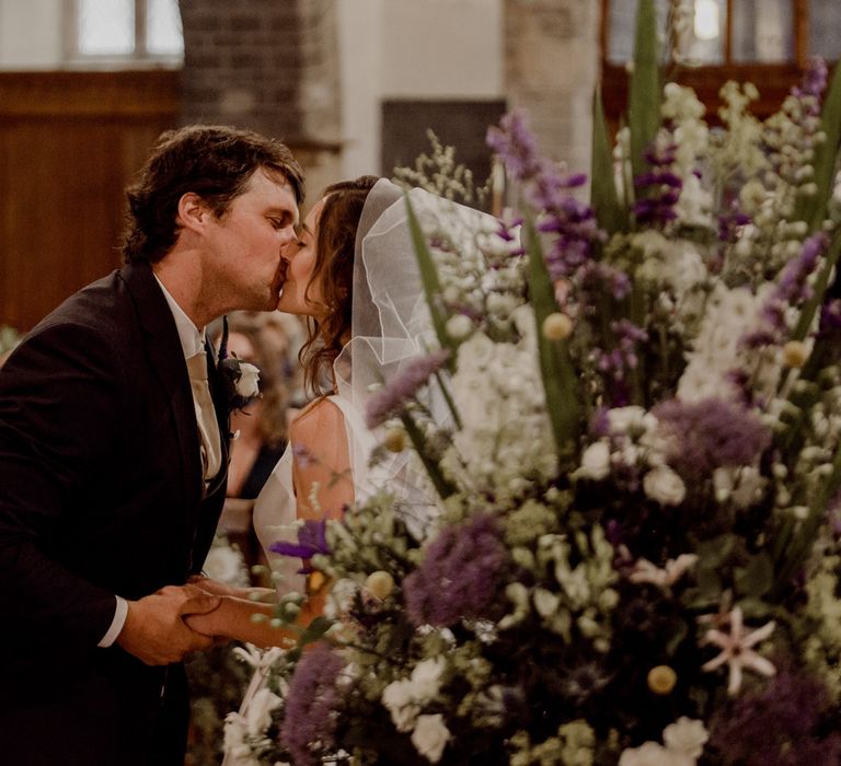 Groom in dark brown suit kisses bride in white Elbeth Gillis wedding dress and veil in church next to white, purple and green florals