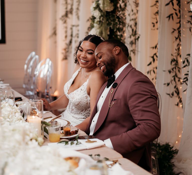 Bride & groom sit together on their wedding day as groom wears deep pink blazer with black bow tie