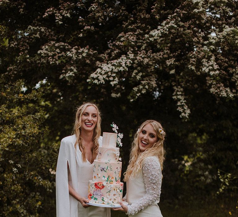 Two brides in a short wedding dress, bridal boots, jumpsuit and cape holding a wedding cake with flower illustration 