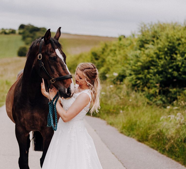 Bride stands with her horse on her wedding day as they walk along the road surrounded by greenery