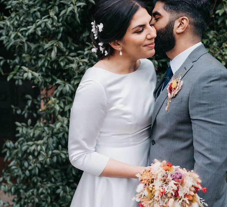 Groom kisses brides cheek as she leans into him whilst holding dried floral bouquet on her wedding day