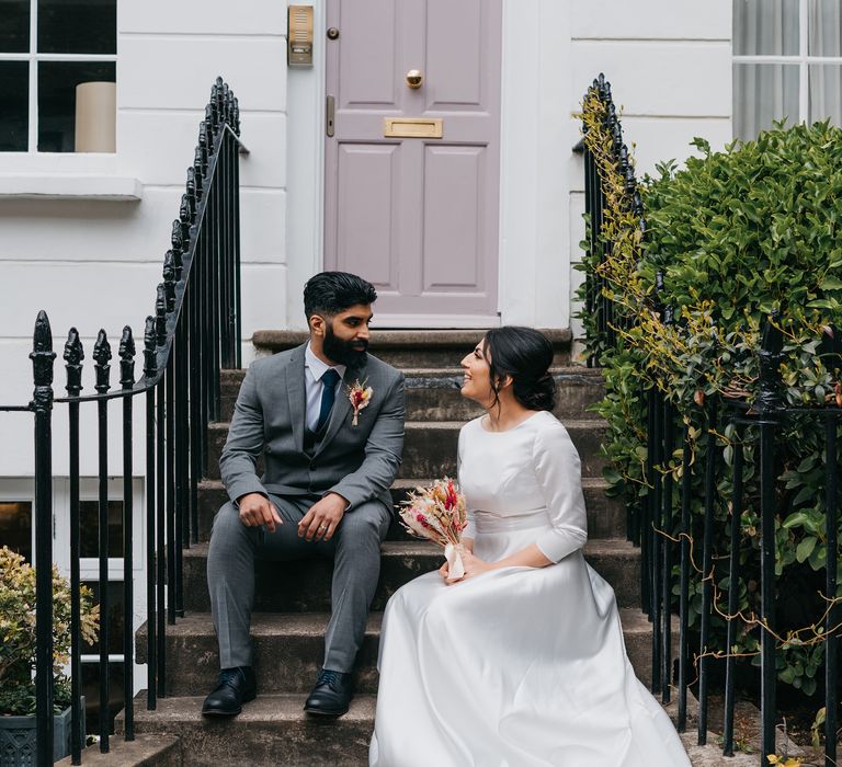 Bride & groom sit on stoop in Chelsea for post-wedding pictures as they look lovingly at one another