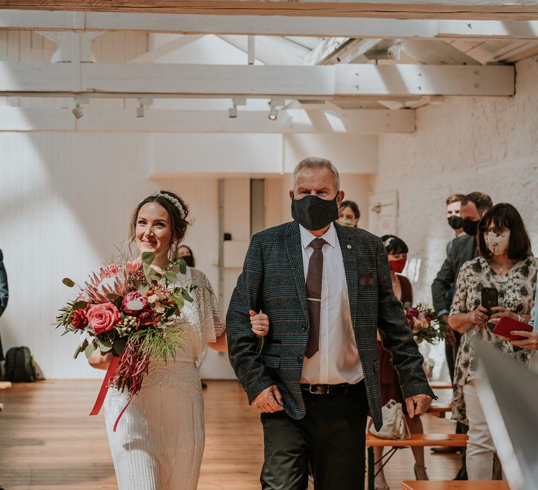 Bride links arms with her father as she walks down the aisle holding brightly coloured bouquet in different tones of reds and pinks 