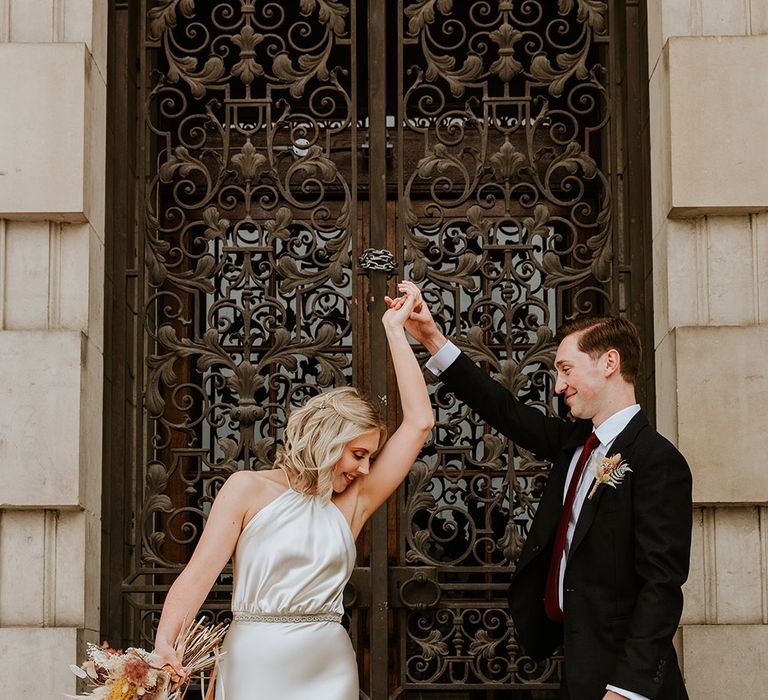 portrait of the groom in a black suit and burgundy tie twirling his bride around in a champagne colour wedding dress as she holds her dried flower bouquet 