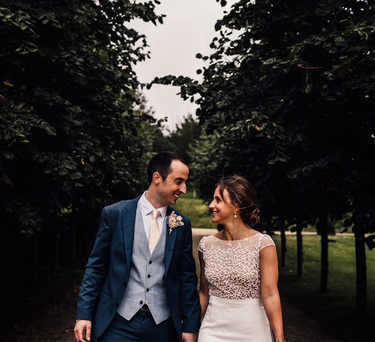 Bride in white Rime Arodaky wedding dress holding dried bridal bouquet walks holding hands with groom in blue three piece suit and dried buttonhole outdoors at summer wedding in Dorset