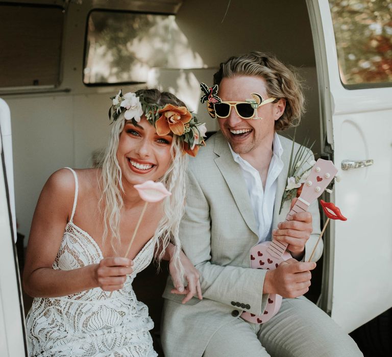 Groom in white shirt and linen suit laughs at smiling bride in cami strap lace boho wedding dress and tropical flower crown as they both sit in the side of a white campervan decorated with tropical flowers for birds of paradise wedding inspiration