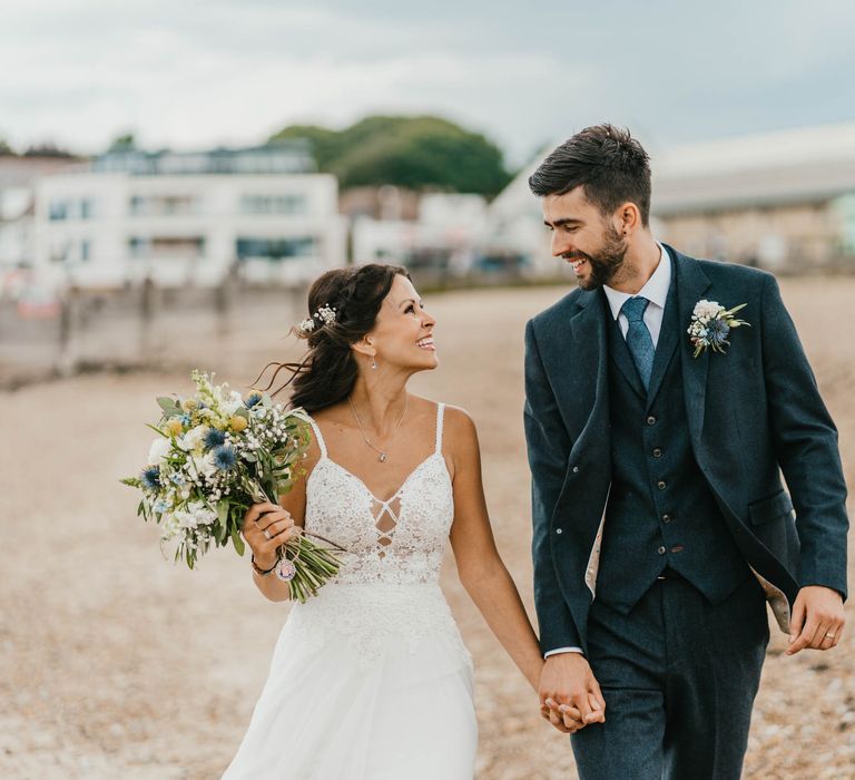 Bride & groom look lovingly at one another as they walk along the beach