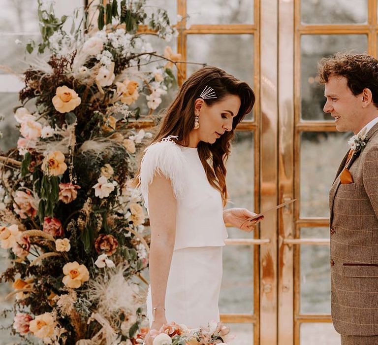 Bride in a fitted wedding dress with feather sleeves reading her vows to her husband in a brown check suit in between orange and yellow vertical flower arrangements 