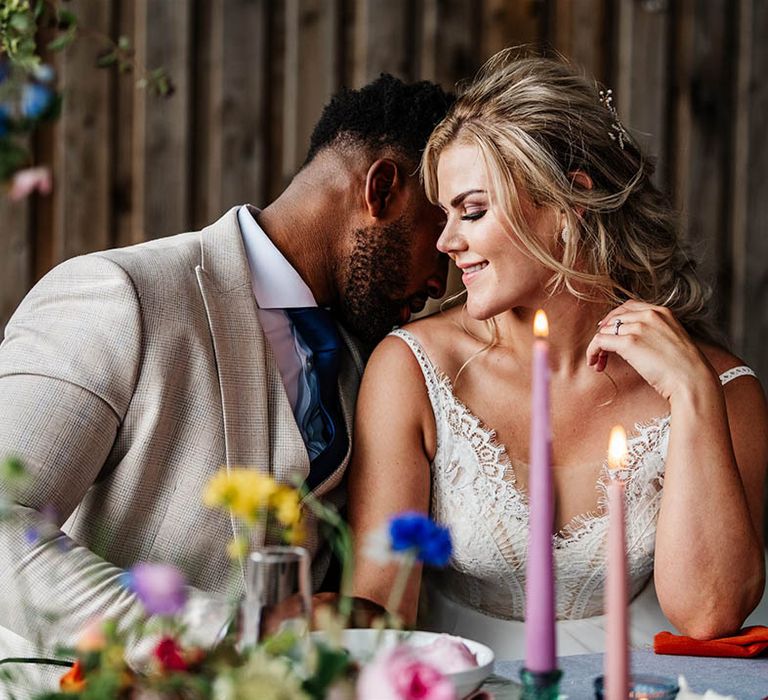 Groom in stone coloured suit and navy tie kissing his brides shoulder at their sweetheart table at Little Wold Vineyard 