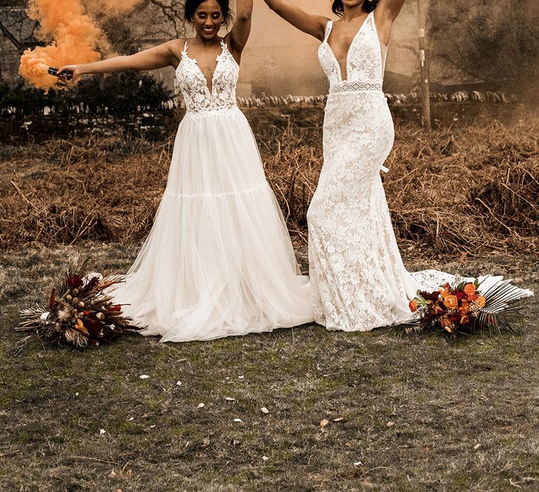 Two brides at at farm wedding in boho wedding dresses holding orange smoke bombs