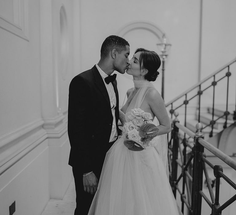 Black and white portrait of a groom in a black tie suit kissing his bride in a romantic tulle wedding dress on the stairs at Clerkenwell House 