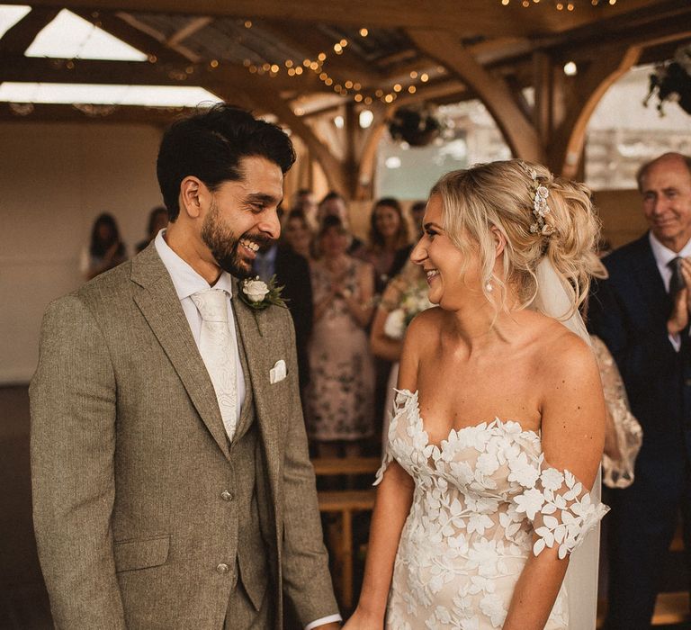 Bride in off the shoulder Enzoani wedding dress and veil smiles with groom in grey three piece suit, white tie and white floral buttonhole during Inkersall Grange Farm wedding ceremony