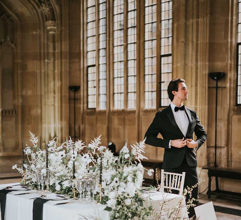 Groom in a tuxedo standing at the intimate reception table decorated with black, white and gold tableware and flowers 