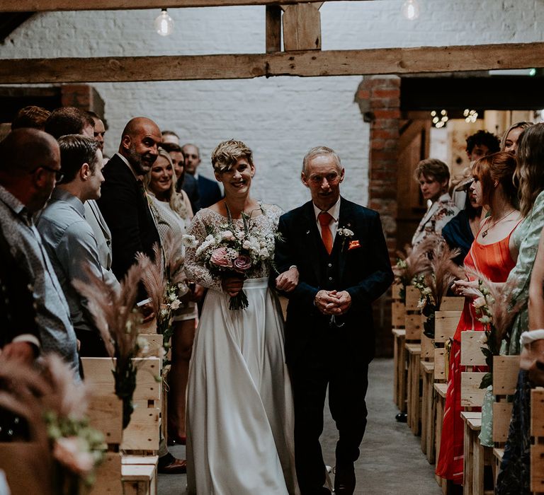 Bride with short hair holding a pink and white wedding bouquet walking down the aisle with her father 