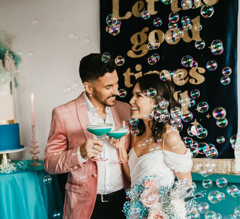 Groom in pink velvet jacket and bride in a cold shoulder wedding dress sipping cocktails with bubbles floating around them 