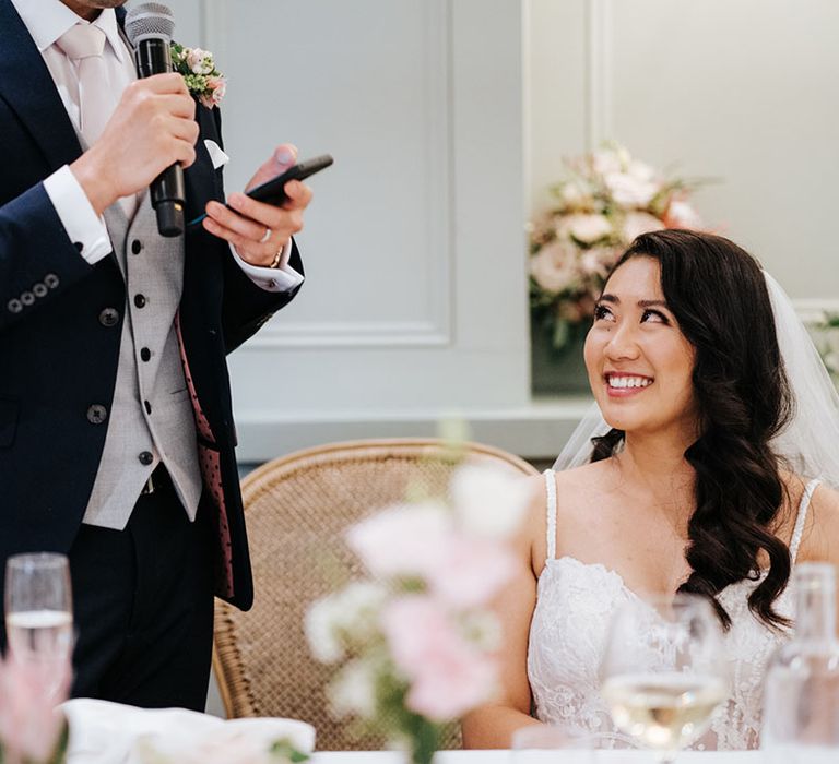 South Asian bride with side swept long curly hair smiling during her grooms wedding speech 