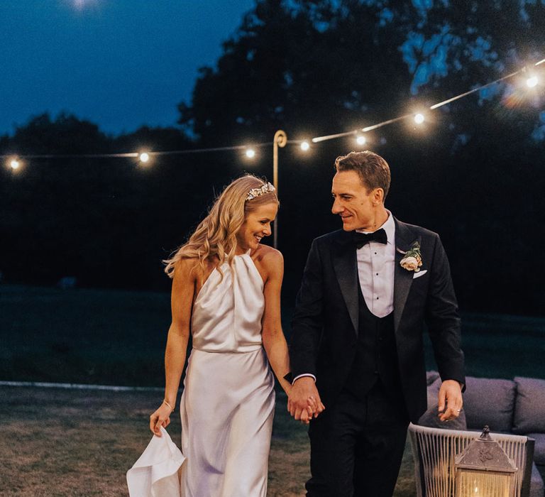 Bride in satin Halfpenny London wedding dress, Jimmy Choo wedding shoes and Victoria Percival headband walks holding hands with groom in black tie and floral buttonhole at sperry tent wedding reception