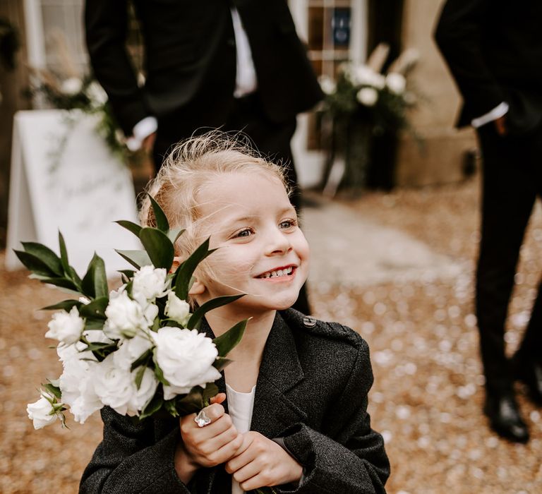 Flower girl in a white dress and black tweed jacket holding a white flower bouquet 