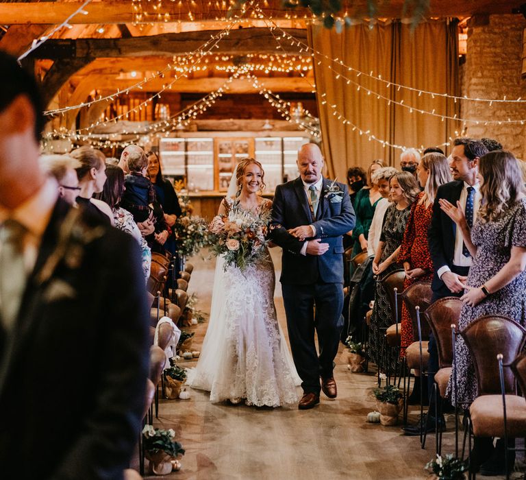 Bride in lace wedding dress with train and veil holding large mixed bridal bouquet walks down aisle with father during rustic barn wedding ceremony