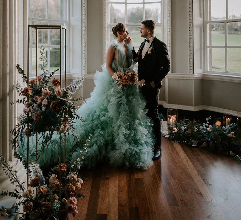 Groom in a tuxedo caressing his brides face as she stands in a dramatic coloured wedding dress at Newburgh Priory surrounded by blush flowers 