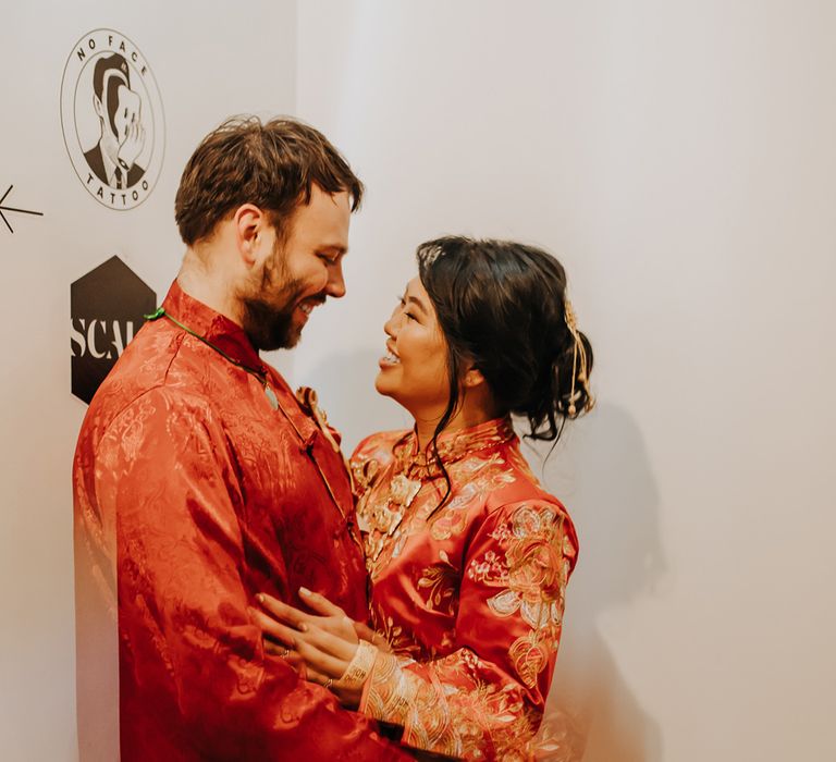 Bride in red and gold traditional Chinese tea ceremony dress stands with groom in black trousers and red traditional Chinese clothing