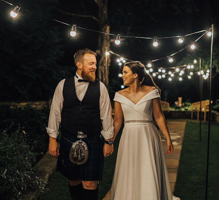 Bride in an off the shoulder wedding dress and groom in a tartan kilt holding hands under a canopy of festoon lights at Foxhill Manor 