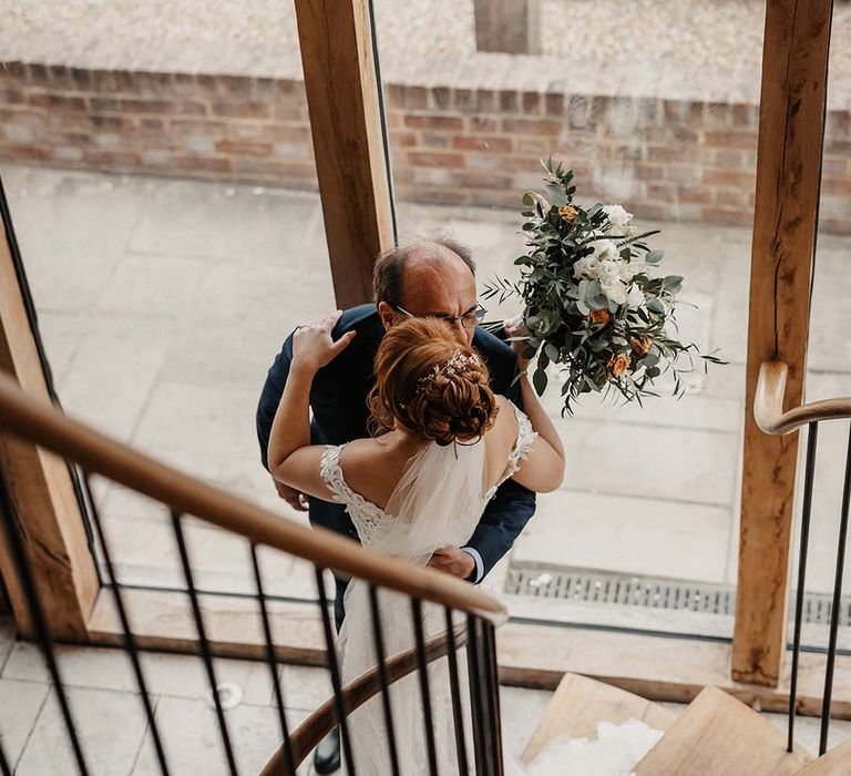 Bride holding white and orange wedding bouquet walks down the stairs to meet her father who wears blue suit