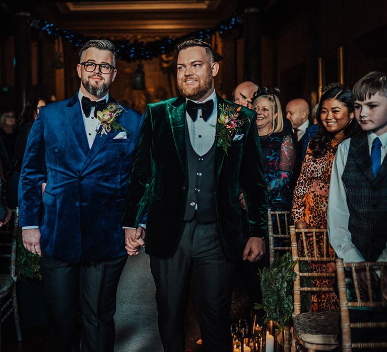 Grooms in blue and green velvet jackets and matching black bow ties walk the aisle together holding hands