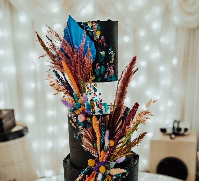 Tall three-tier wedding cake with dark frosting, white ribbon and colourful dried flower decoration