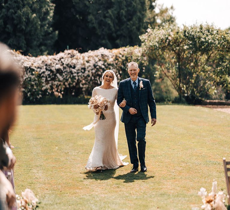 Father of the bride in checkered blue suit walks the bride in beaded wedding dress down the aisle for outdoor wedding ceremon